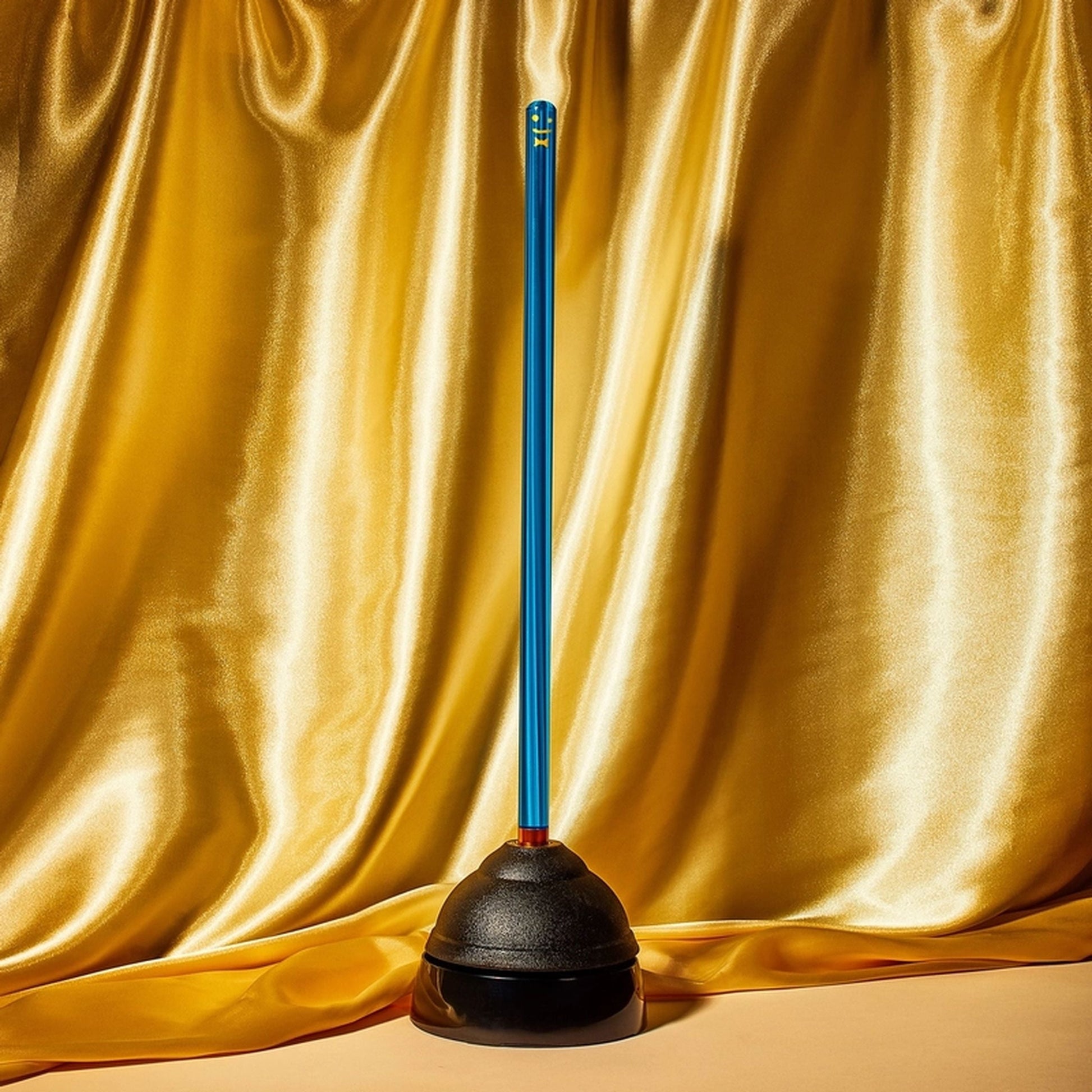 Staff The Plunger by Staff features a blue handle and stands upright with a sanitary drip tray, shown on a smooth surface against a shiny gold satin fabric backdrop.