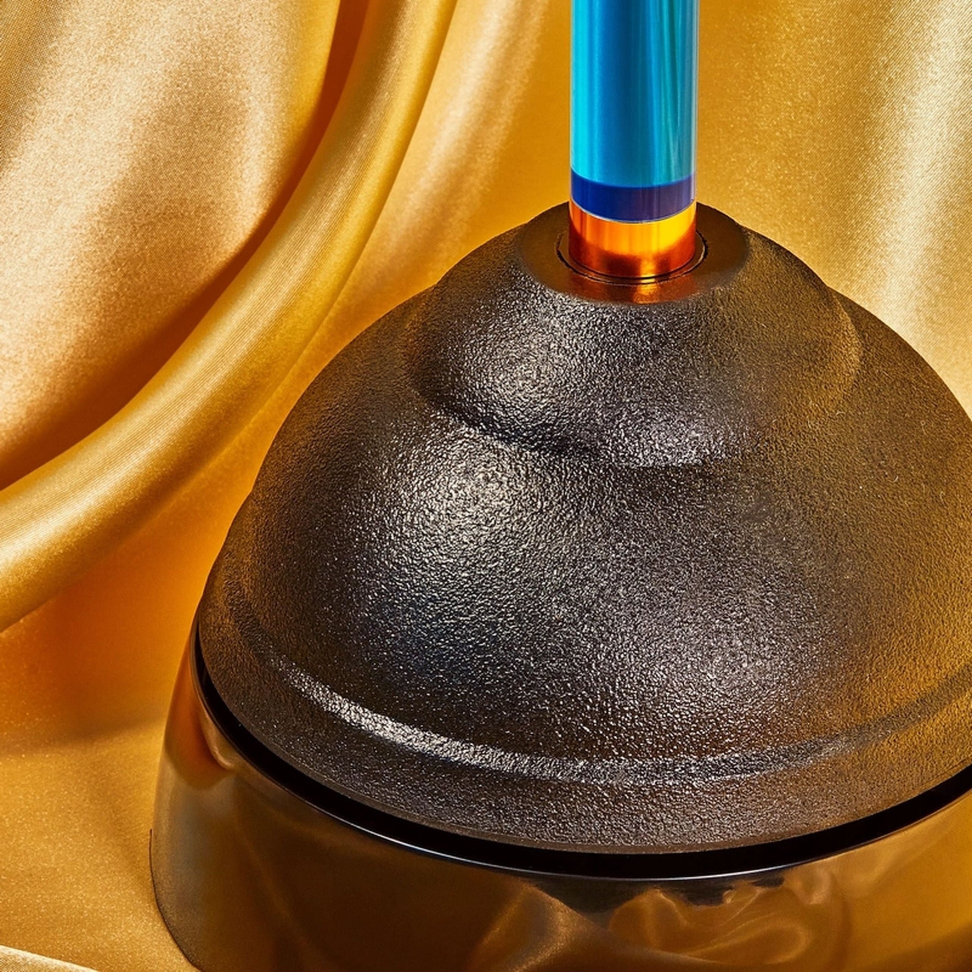 A close-up of Staff The Plunger, a bathroom essential by Staff with a blue and orange handle, set against shiny gold satin fabric.
