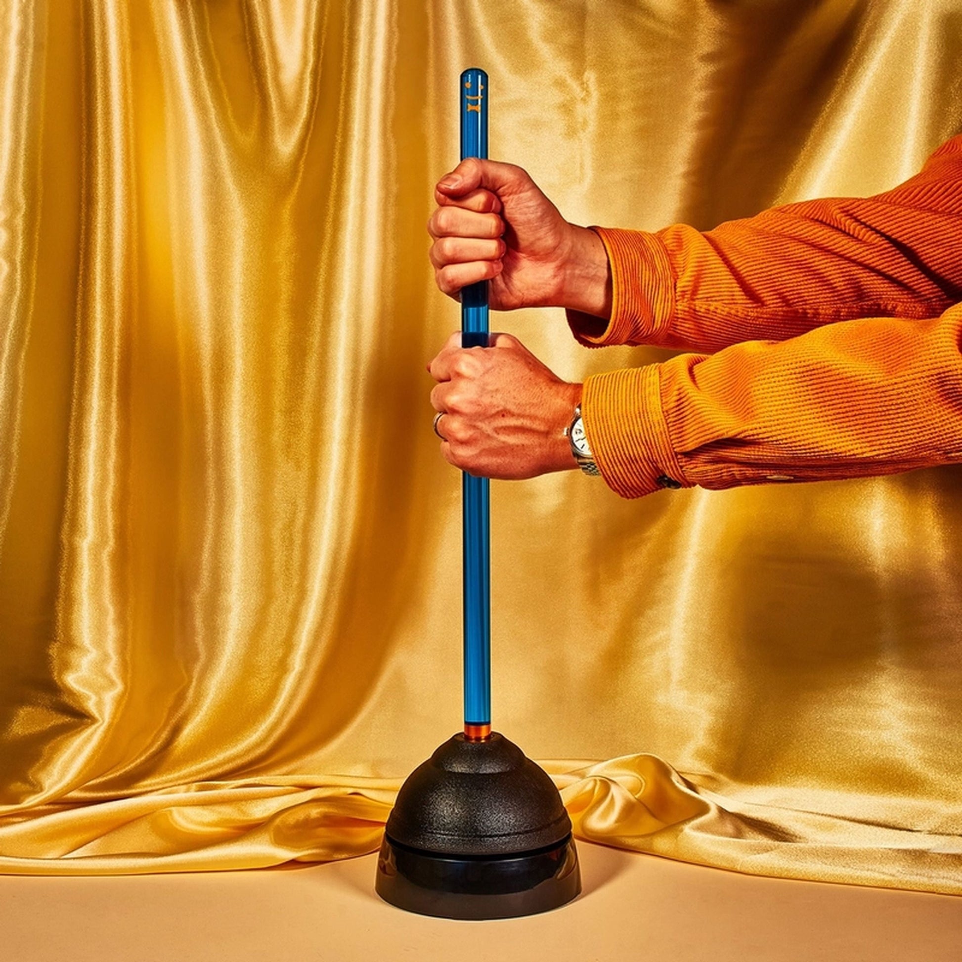 Against a gold satin backdrop, hands and forearms hold Staff The Plunger by Staff, featuring a blue handle. The person wears an orange shirt; the scene is brightly lit.