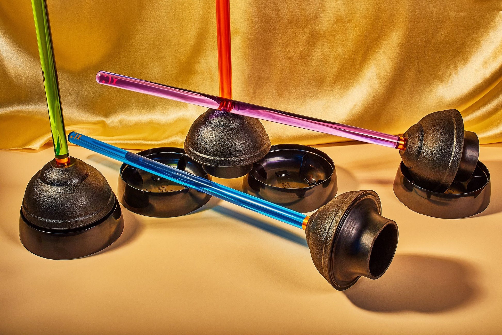 Staff The Plunger by Staff—a colorful plastic plunger with a black rubber cup and vibrant handle—is showcased on a beige surface against gold fabric, arranged in diagonal and horizontal positions.