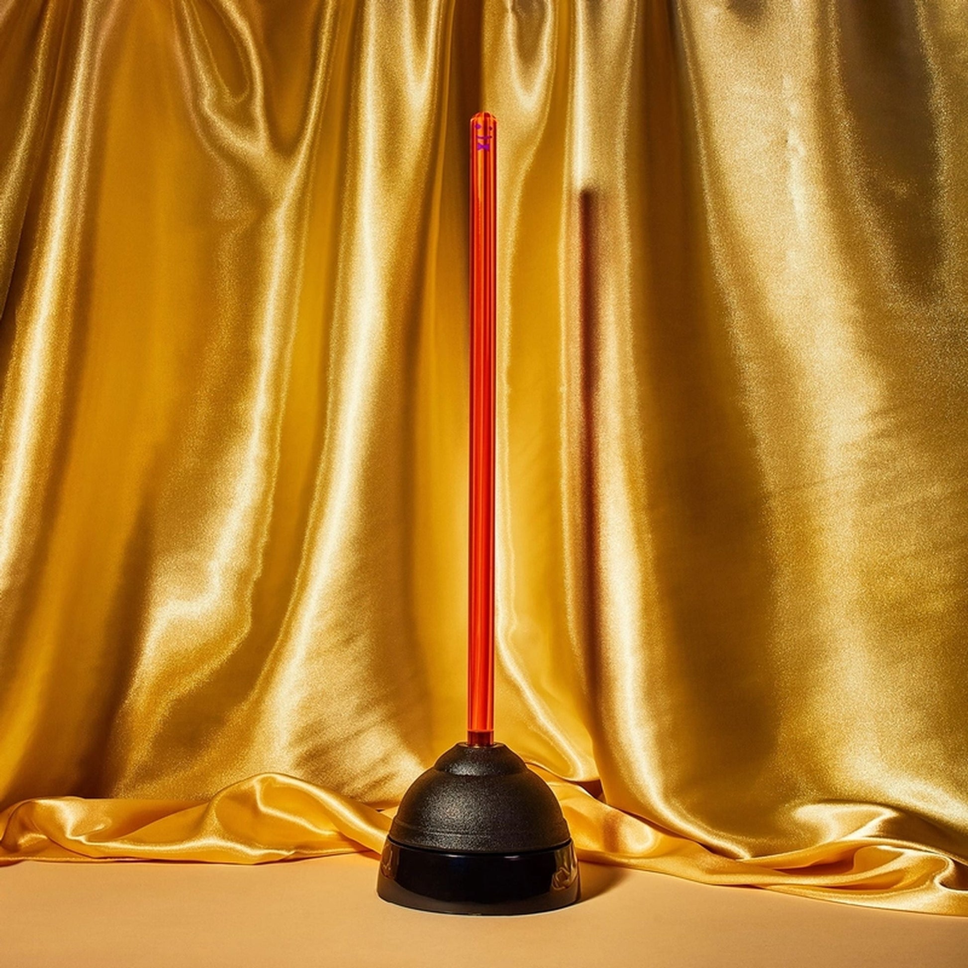 Staff The Plunger by Staff is a black toilet plunger with a vibrant red handle, shown upright on a sanitary drip tray before gold satin fabric—a must-have bathroom essential.
