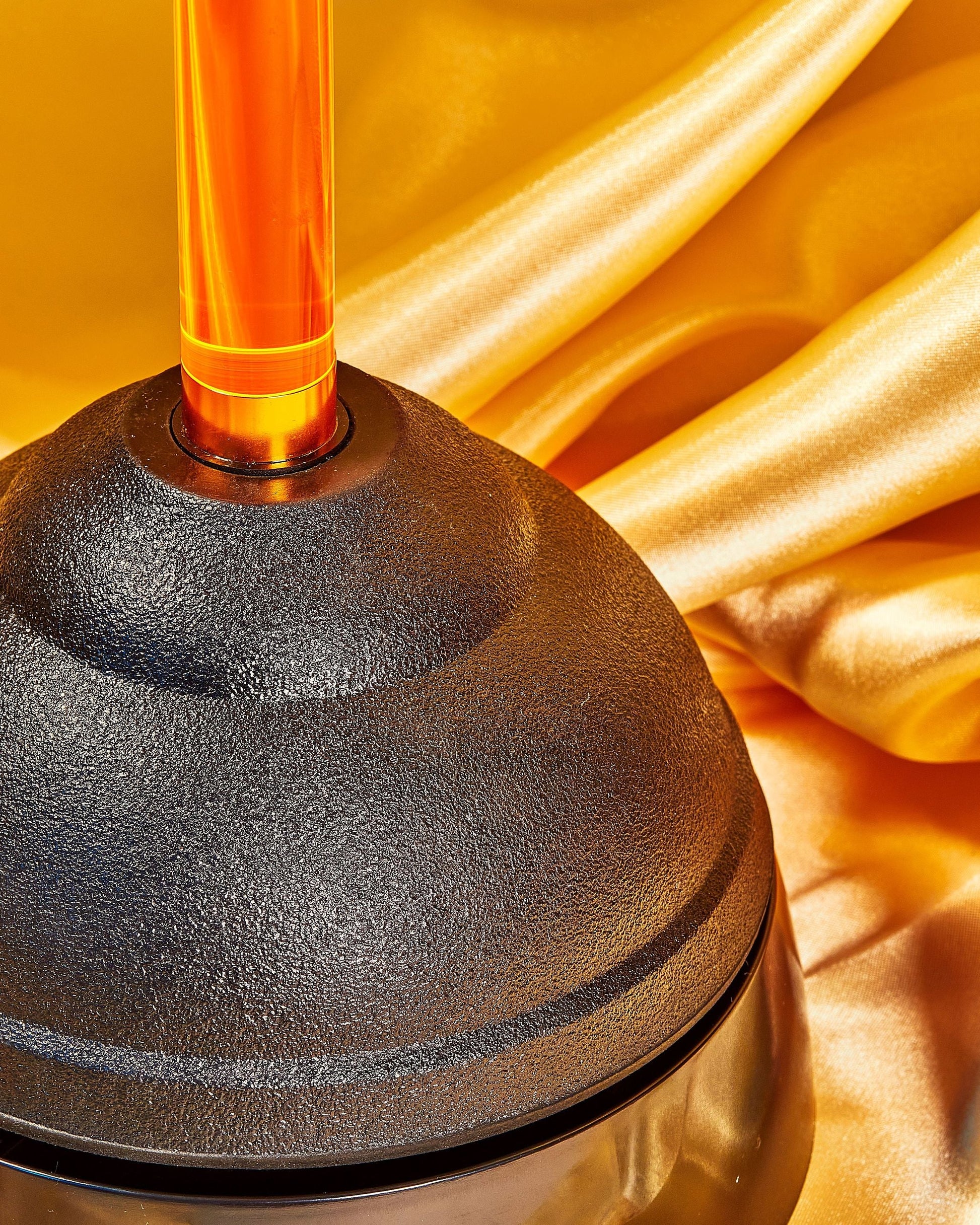 A bathroom essential, Staff The Plunger by Staff, featuring a black cup and orange handle, rests on shiny gold satin fabric with graceful folds and a glossy finish.
