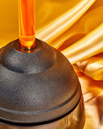 A bathroom essential, Staff The Plunger by Staff, featuring a black cup and orange handle, rests on shiny gold satin fabric with graceful folds and a glossy finish.