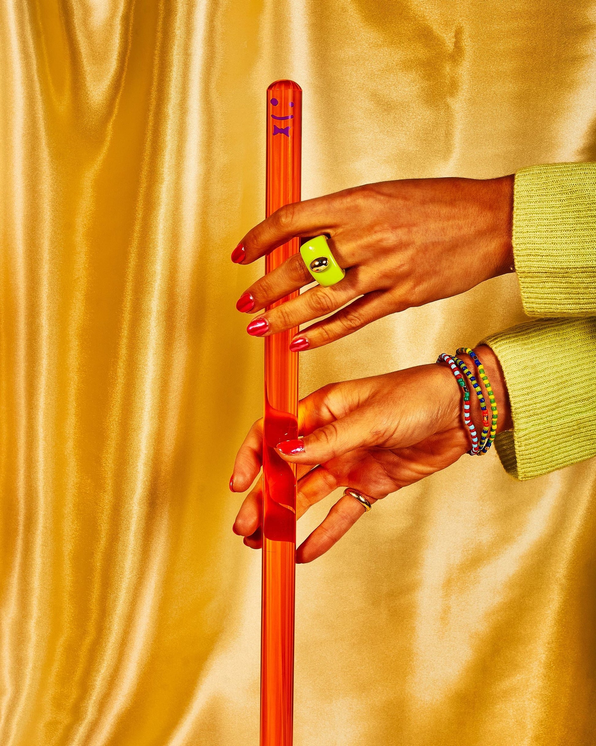 Two hands with red nails and colorful jewelry hold Staff The Plunger—a translucent orange rod by Staff—against shiny gold fabric. One hand wears a green smiley face ring, turning this bathroom essential into a chic accessory.