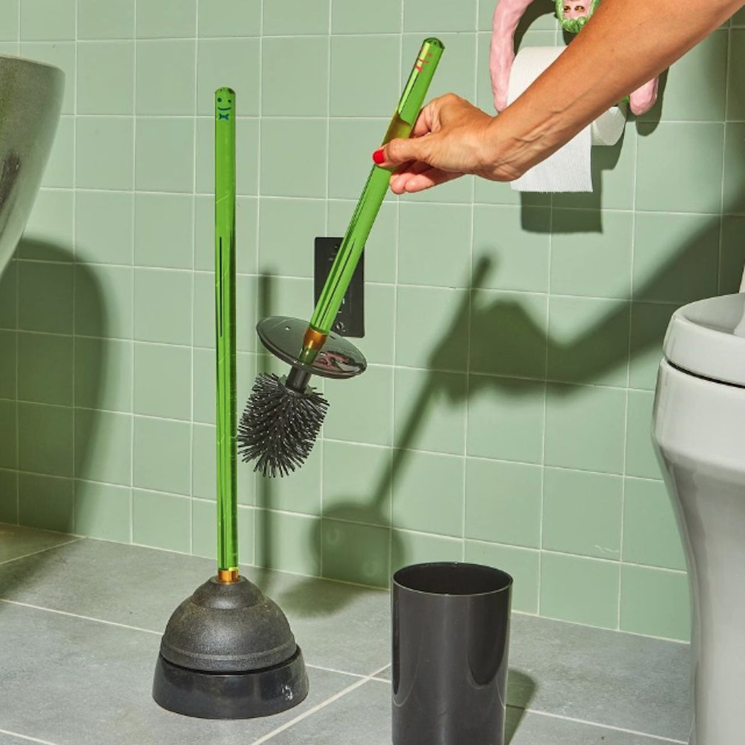 A hand grabs the Staff The Toilet Brush - Green by Staff from its holder next to a matching plunger in a tiled bathroom, with a toilet and toilet paper roll visible in the background.