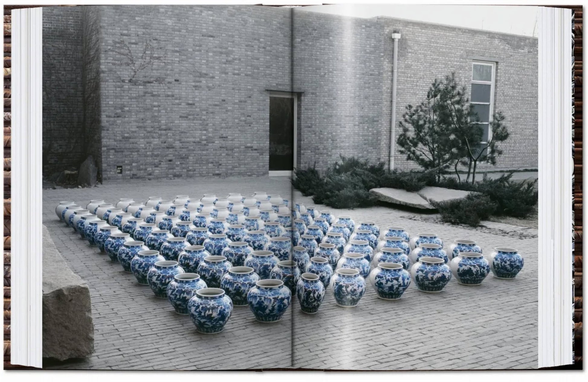 Blue and white porcelain jars, like those in "TASCHEN Ai Weiwei" by TASCHEN, are arranged in a grid on a brick courtyard beside a modern brick building with a tree and shrubs nearby.