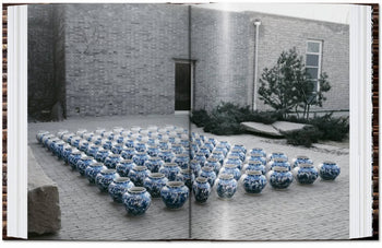 Blue and white porcelain jars, like those in "TASCHEN Ai Weiwei" by TASCHEN, are arranged in a grid on a brick courtyard beside a modern brick building with a tree and shrubs nearby.