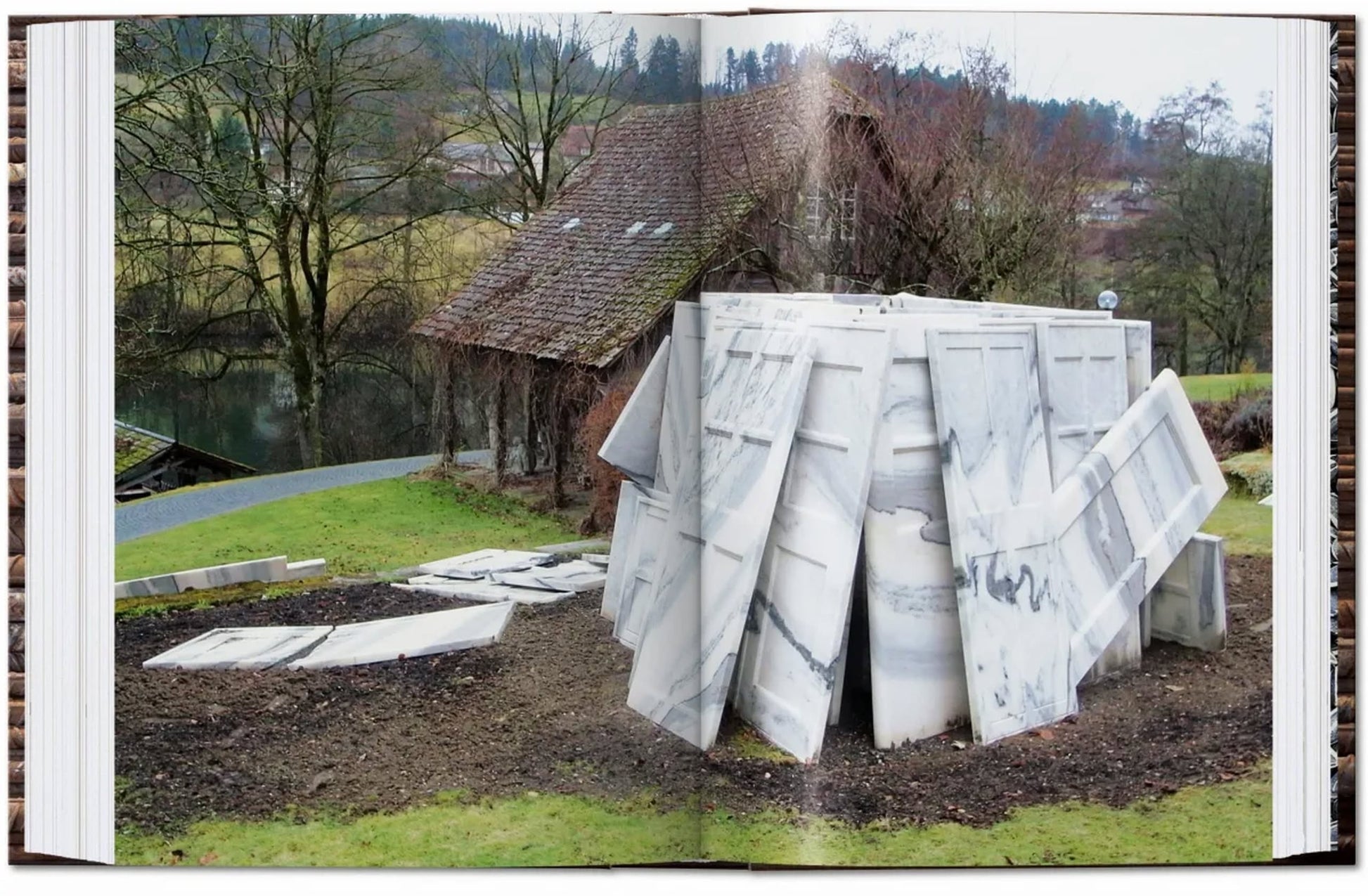 Large, leaning marble slabs stand on grass near a rustic building with bare trees and a pond, evoking themes from the TASCHEN Ai Weiwei monograph by TASCHEN. Scattered marble pieces surround the sculpture in this rural scene.