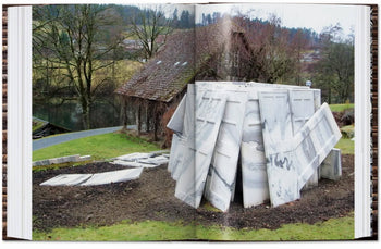 Large, leaning marble slabs stand on grass near a rustic building with bare trees and a pond, evoking themes from the TASCHEN Ai Weiwei monograph by TASCHEN. Scattered marble pieces surround the sculpture in this rural scene.