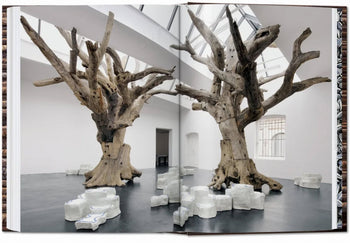 A striking image from the TASCHEN Ai Weiwei monograph shows two large leafless tree sculptures of weathered wood, surrounded by white abstract ceramics on a dark floor beneath a skylight, spanning two pages.