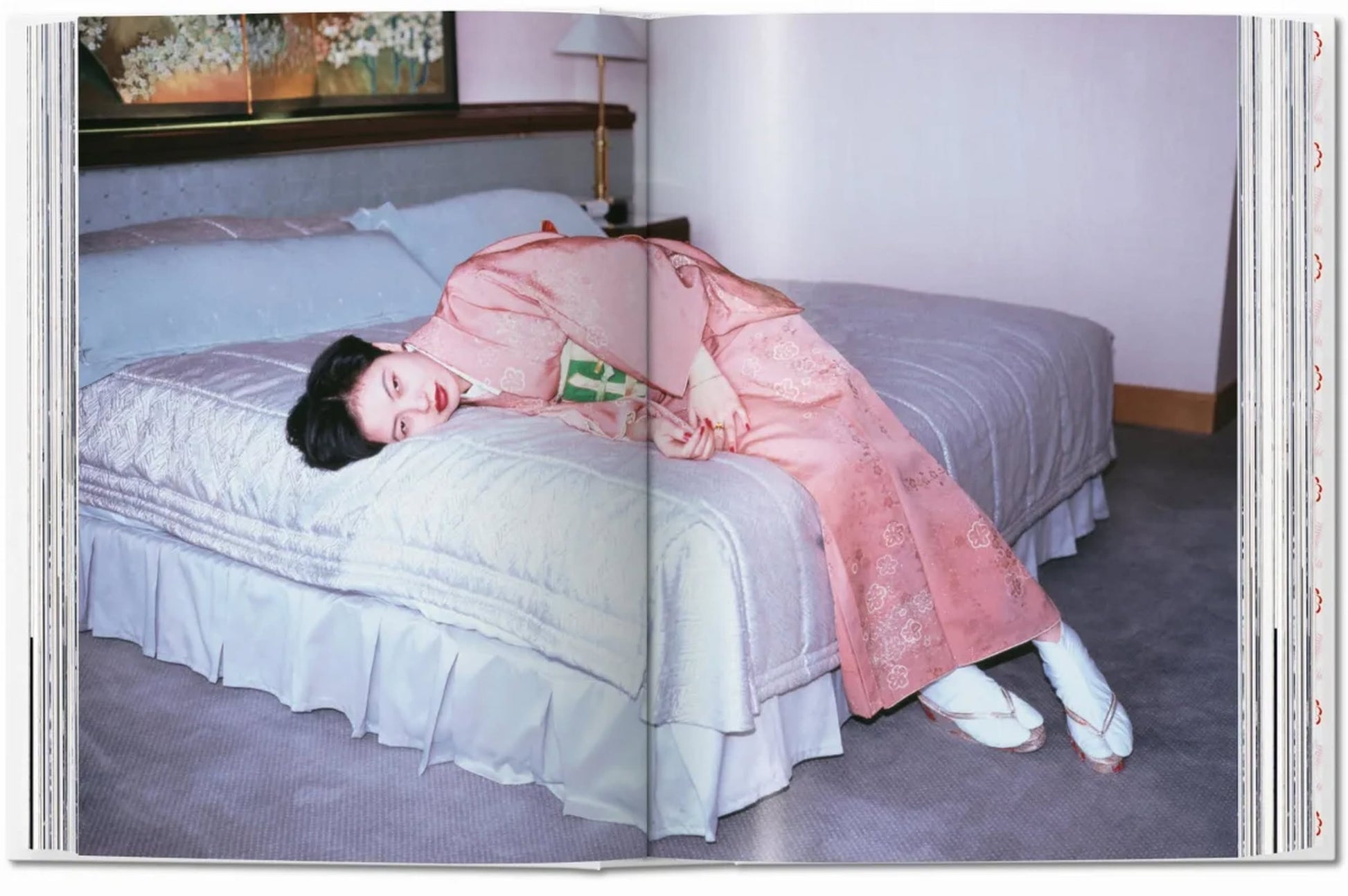 A person in a pink kimono and white ballet shoes lies sideways on a tidy bed, holding a book, radiating serenity in soft light—an intimate scene reminiscent of Araki’s style, inspired by TASCHEN Araki from TASCHEN.