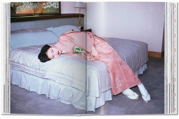 A person in a pink kimono and white ballet shoes lies sideways on a tidy bed, holding a book, radiating serenity in soft light—an intimate scene reminiscent of Araki’s style, inspired by TASCHEN Araki from TASCHEN.