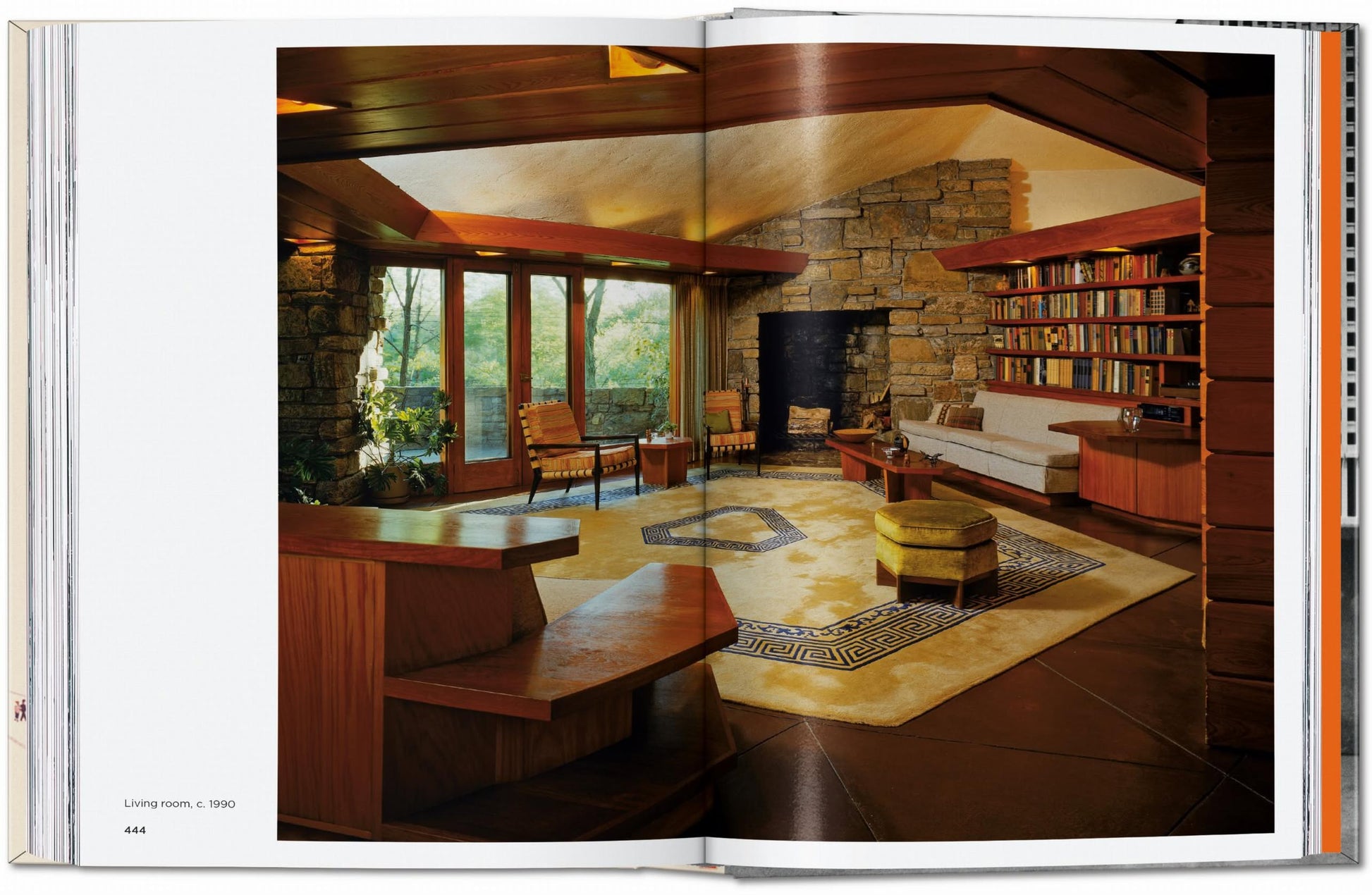 A photo of a spacious living room showcases stone walls, wood accents, built-in shelves with TASCHEN Frank Lloyd Wright books, large windows, a fireplace, and mid-century modern furniture on a patterned rug.