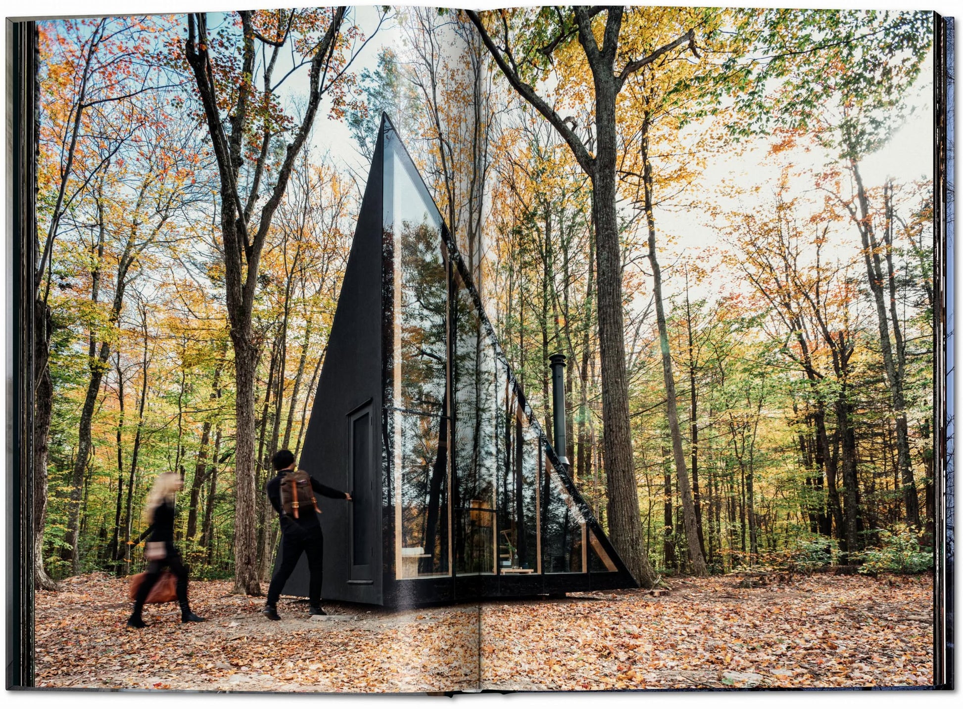 Two people approach a striking black A-frame cabin with large glass windows—an example from TASCHEN Homes for Our Time: Small Houses (XL)—nestled among colorful autumn trees.