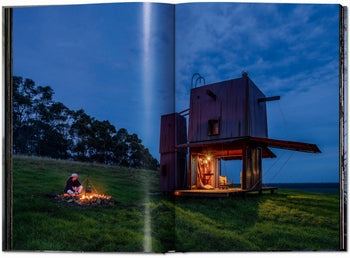 A person sits by a campfire near a modern, boxy cabin—petite architecture featured in TASCHEN Homes for Our Time: Small Houses (XL)—warmly lit on a grassy field at dusk under a cloudy sky.