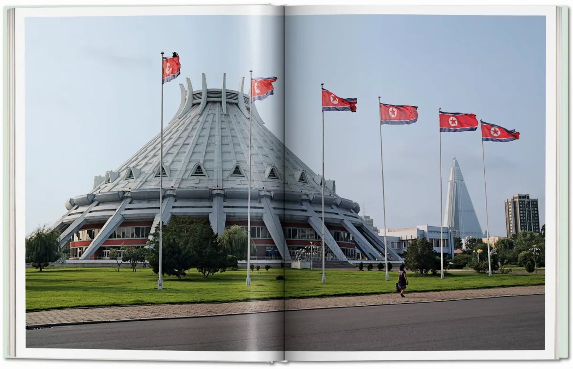 A large, futuristic dome rises behind North Korean flags, echoing scenes from TASCHENs Inside North Korea, as seen across a broad road bordered by greenery.