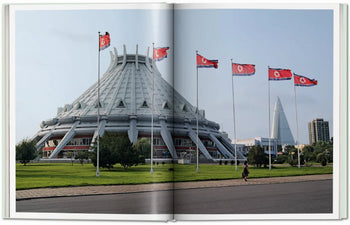 A large, futuristic dome rises behind North Korean flags, echoing scenes from TASCHENs Inside North Korea, as seen across a broad road bordered by greenery.