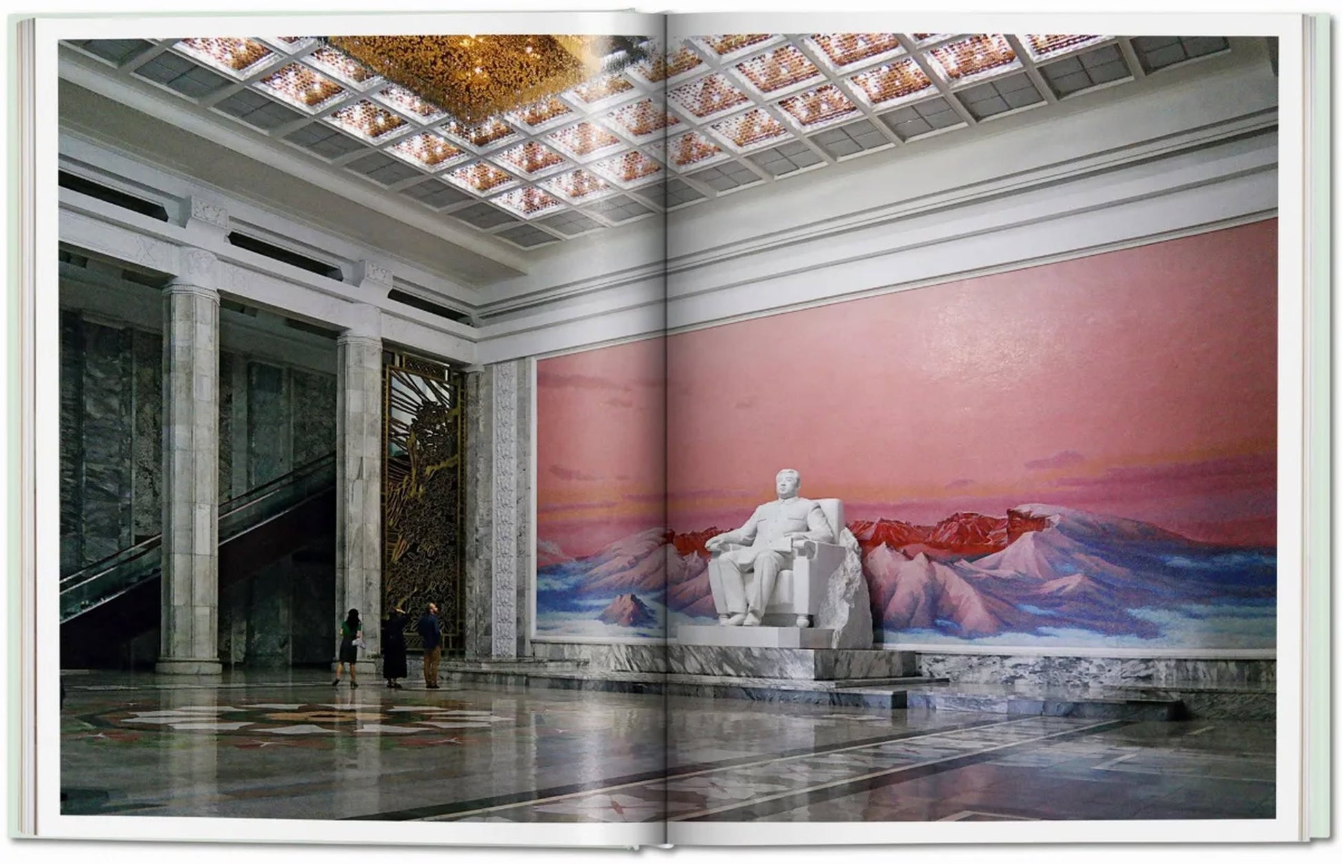 A grand marble hall, reminiscent of Oliver Wainwright’s imagery in TASCHEN Inside North Korea, features tall columns, a white statue before a pink mountain mural, two admirers, and an escalator from TASCHEN.