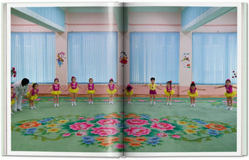 A group of young children in colorful costumes bow in a vibrant, pastel room with floral carpet and cartoon decor—an inviting setting reminiscent of scenes from TASCHEN Inside North Korea by TASCHEN; an adult stands on the left.