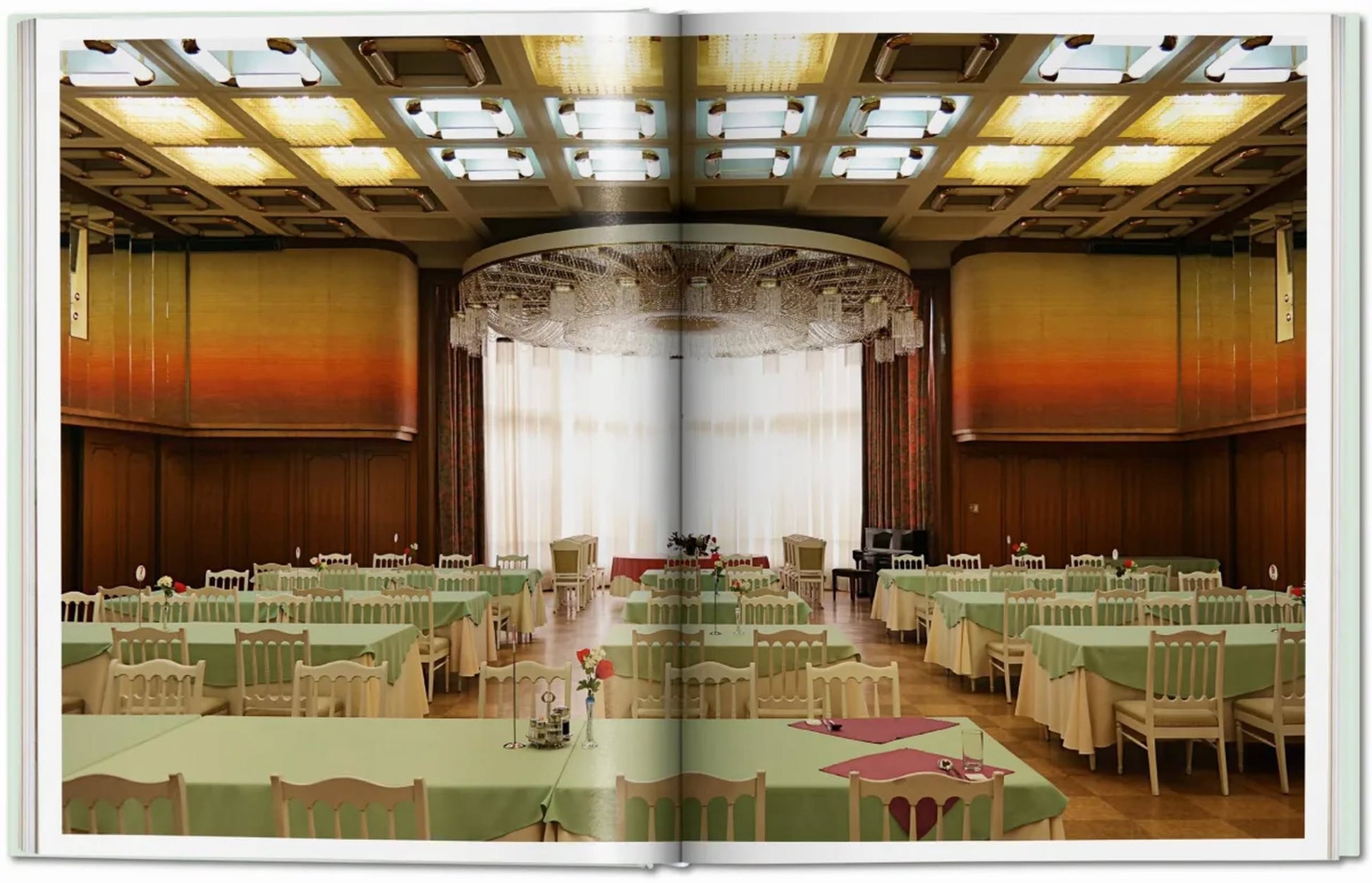 A photo of a grand banquet hall with green tablecloths, white chairs, a curtained stage, and ornate ceiling spans the pages of TASCHEN Inside North Korea by TASCHEN.