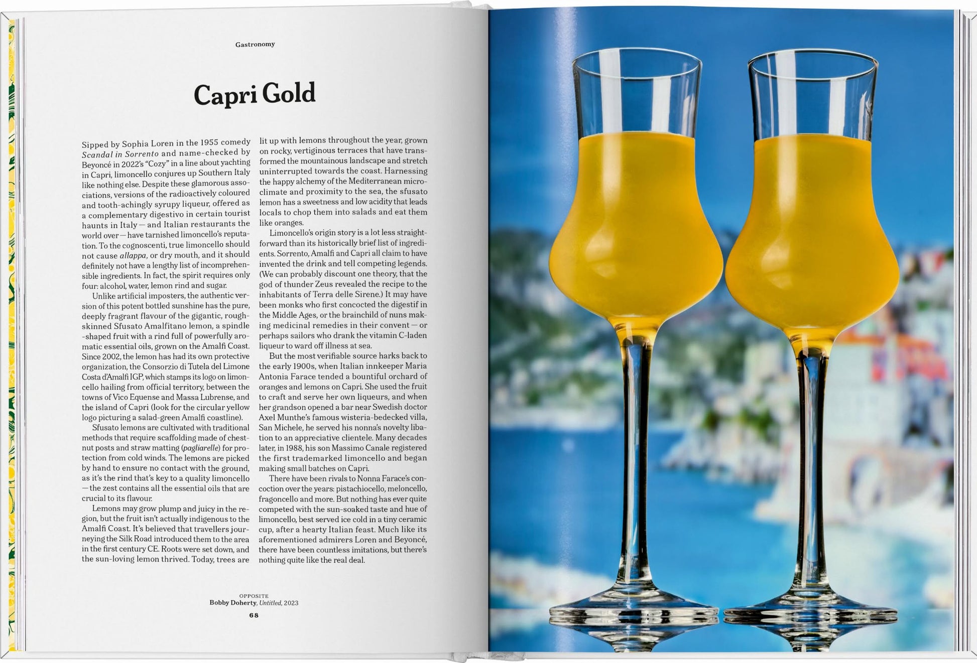 An open TASCHEN The Gourmands Lemon book displays recipes on the left, while two glasses of lemon liqueur sit on the right, with a blurred seaside town in the background.