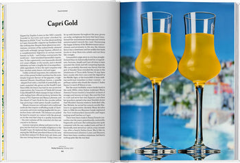 An open TASCHEN The Gourmands Lemon book displays recipes on the left, while two glasses of lemon liqueur sit on the right, with a blurred seaside town in the background.