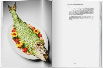 On the left page of TASCHEN The Gourmands Lemon: A Collection of Stories and Recipes, a whole salmon is artfully plated with cucumber scales, lemon slices, shrimp, and tomato flowers on a white oval dish.
