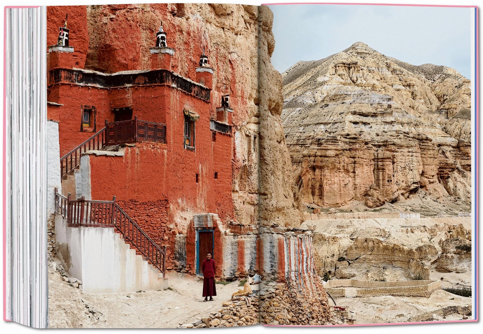 A person in red robes stands outside a red and white cliffside monastery amid rugged mountains—a scene worthy of the TASCHEN The New York Times Explorer: 100 Dream Trips Around the World.