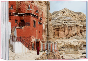 A person in red robes stands outside a red and white cliffside monastery amid rugged mountains—a scene worthy of the TASCHEN The New York Times Explorer: 100 Dream Trips Around the World.