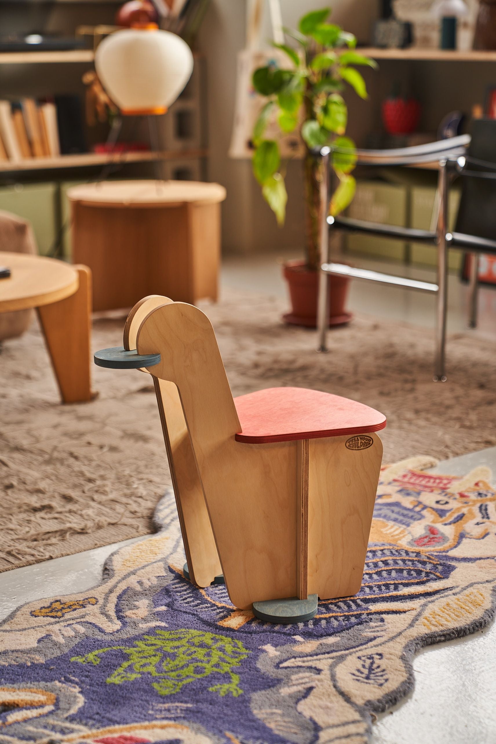 The Tell Your Children x Snug Woodwork The Seating Duck, a playful bird-shaped wooden chair with a red seat and gray footrest, stands on a colorful rug in a cozy modern living room with plants and wooden furniture.