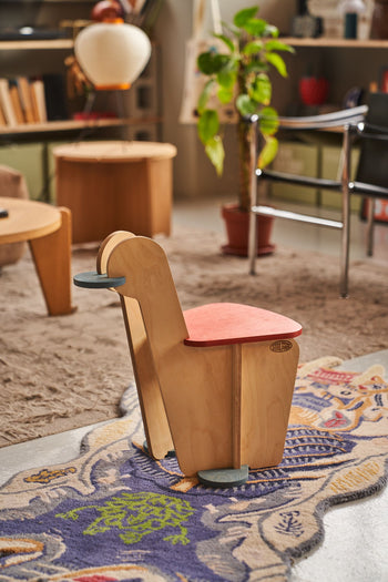 The Tell Your Children x Snug Woodwork The Seating Duck, a playful bird-shaped wooden chair with a red seat and gray footrest, stands on a colorful rug in a cozy modern living room with plants and wooden furniture.
