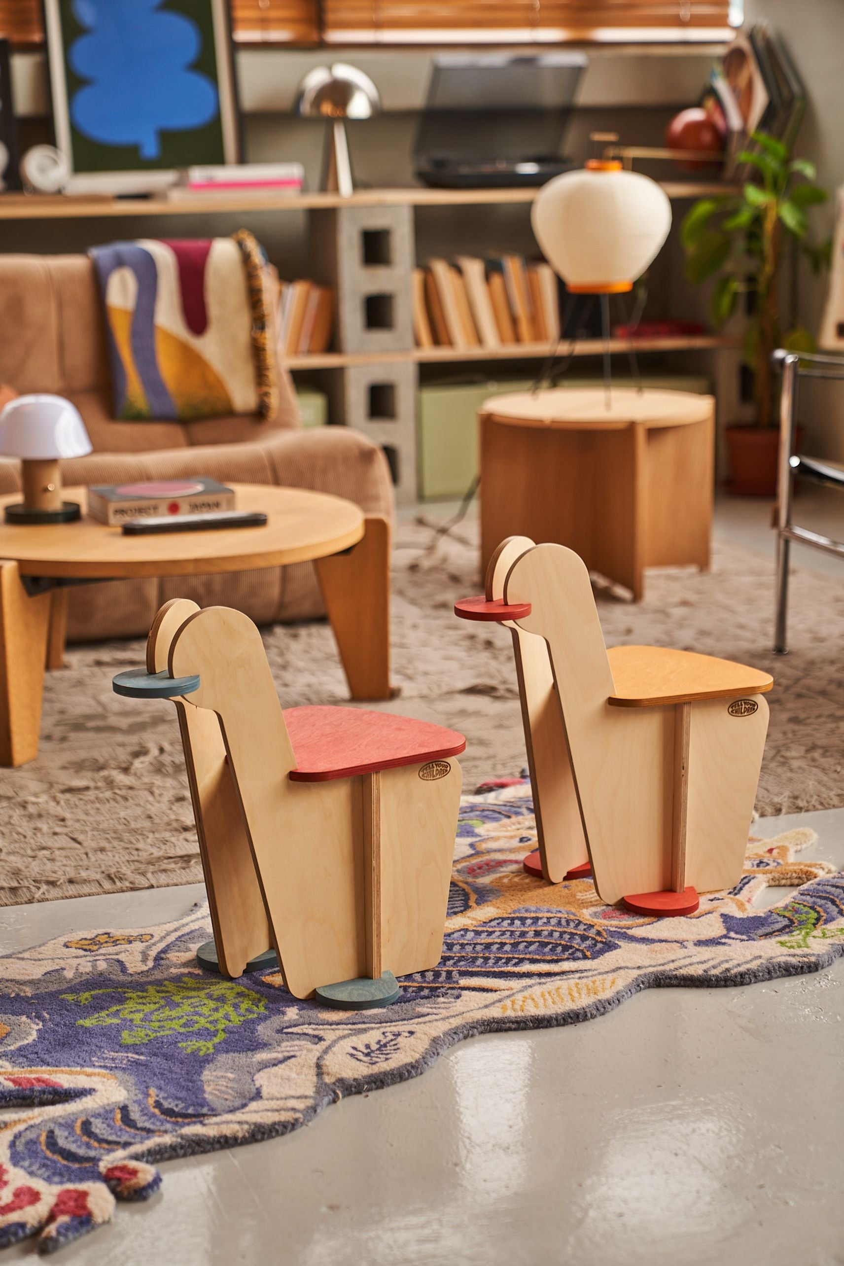 Two modern wooden chairs with colorful seat and arm accents sit on a decorative rug in a stylish living room with mid-century furniture, books, plants, and playful Tell Your Children x Snug Woodwork The Seating Duck rockers in the background.
