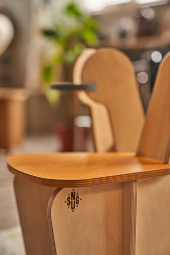 A close-up of the Tell Your Children x Snug Woodwork The Seating Duck chair shows its smooth, curved edges and light wood finish, with a small engraved SNG logo on the side against a softly blurred background. Inspired by playful playground rockers.