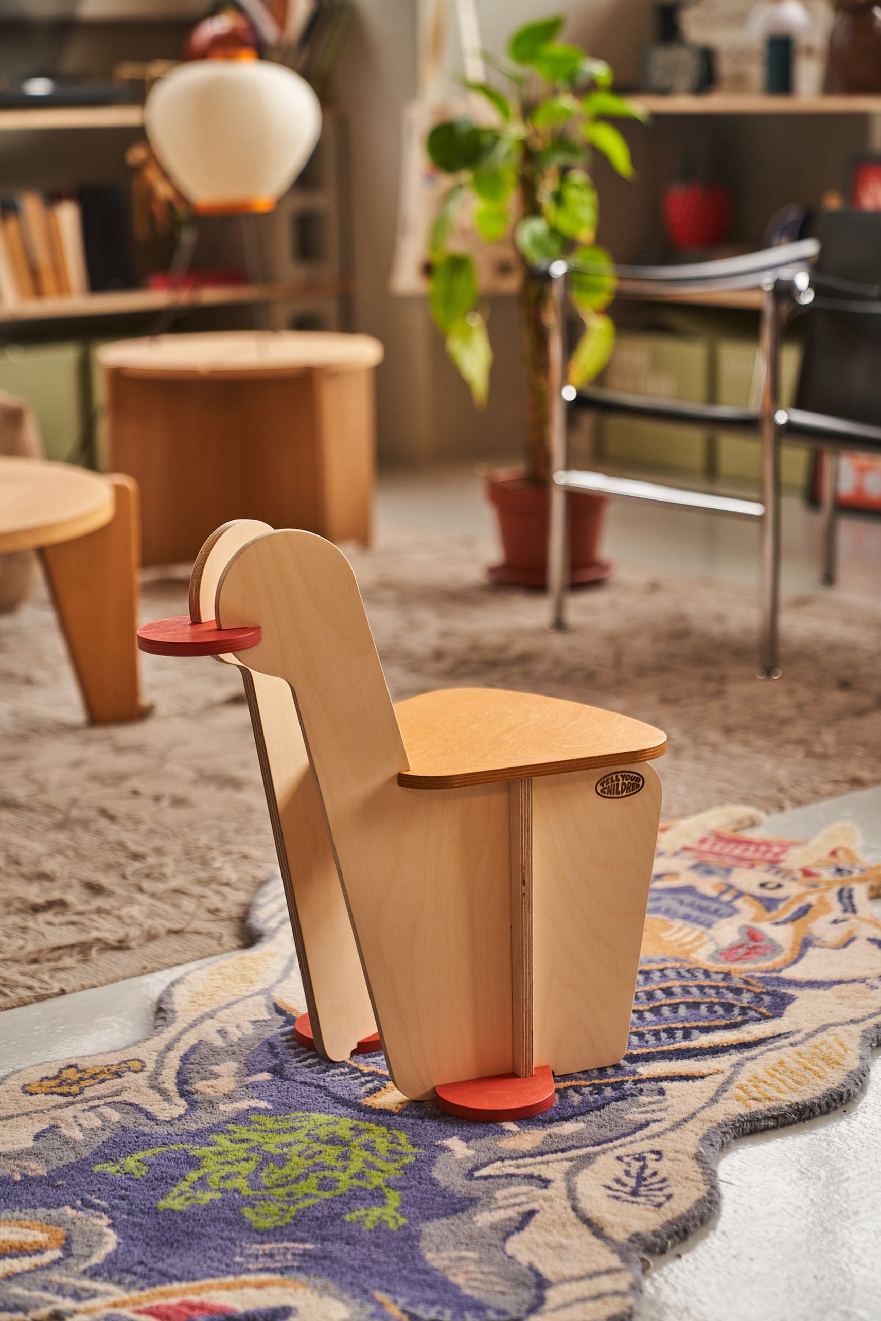 The Tell Your Children x Snug Woodwork The Seating Duck, a small modern wooden chair with red circular feet and armrests inspired by playground rockers, sits on a patterned rug in a cozy, plant-filled living room.