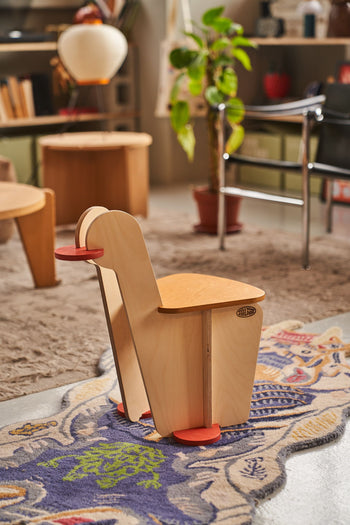 The Tell Your Children x Snug Woodwork The Seating Duck, a small modern wooden chair with red circular feet and armrests inspired by playground rockers, sits on a patterned rug in a cozy, plant-filled living room.