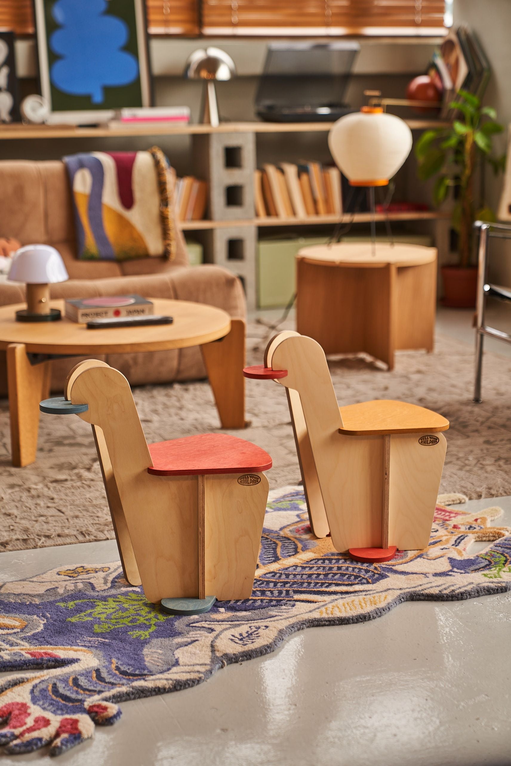 Two vibrant Tell Your Children x Snug Woodwork The Seating Duck chairs with curved backs sit on a patterned rug in a cozy, modern living room featuring a tan sofa, wooden tables, books, and artwork in the background.