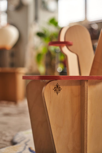 Close-up of the Tell Your Children x Snug Woodwork The Seating Duck chair, crafted in wood with red trim, engraved SNG letters and a compass. A softly blurred backdrop reveals indoor plants, furniture, and playful playground rockers.