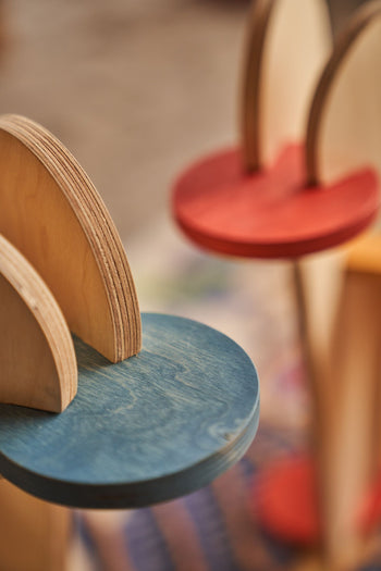 Close-up of Tell Your Children x Snug Woodwork The Seating Duck—colorful, round wooden shelves with blue and red surfaces and vertical supports—displayed indoors with a blurred background, evoking playful playground rockers.