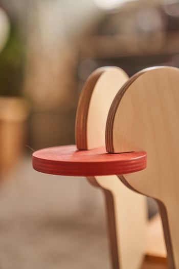 Close-up of Tell Your Children x Snug Woodwork The Seating Duck, a wooden duck-shaped seat with a red circular tongue between two pale wood layers forming the head. Blurred background.