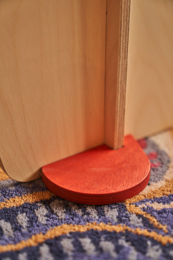 A close-up of Tell Your Children x Snug Woodwork The Seating Duck shows two light wooden boards joined at a right angle, supported by a red, semi-circular wooden piece on a blue and yellow patterned carpet.