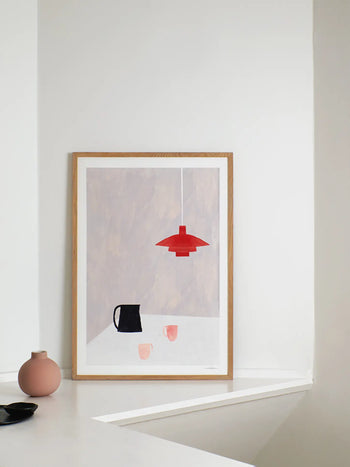 The Poster Club Ana Frois - Orange Pendant (30x40) giclée print features a minimalist table scene with black pitcher, pink cups, and red pendant lamp—framed and displayed beside a pink vase and black dish.