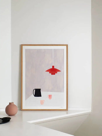 The Poster Clubs Ana Frois - Orange Pendant (50x70) print rests against a white wall, depicting a black pitcher, two pink cups, and an orange pendant lamp. A small pink vase and dark dish are placed nearby on the white surface.