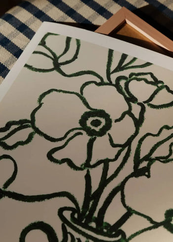A close-up of The Poster Club Anna Johansson - Monday (30x40), featuring a dark green line drawing of flowers and leaves on white paper, placed on checkered fabric with part of a wooden frame visible.
