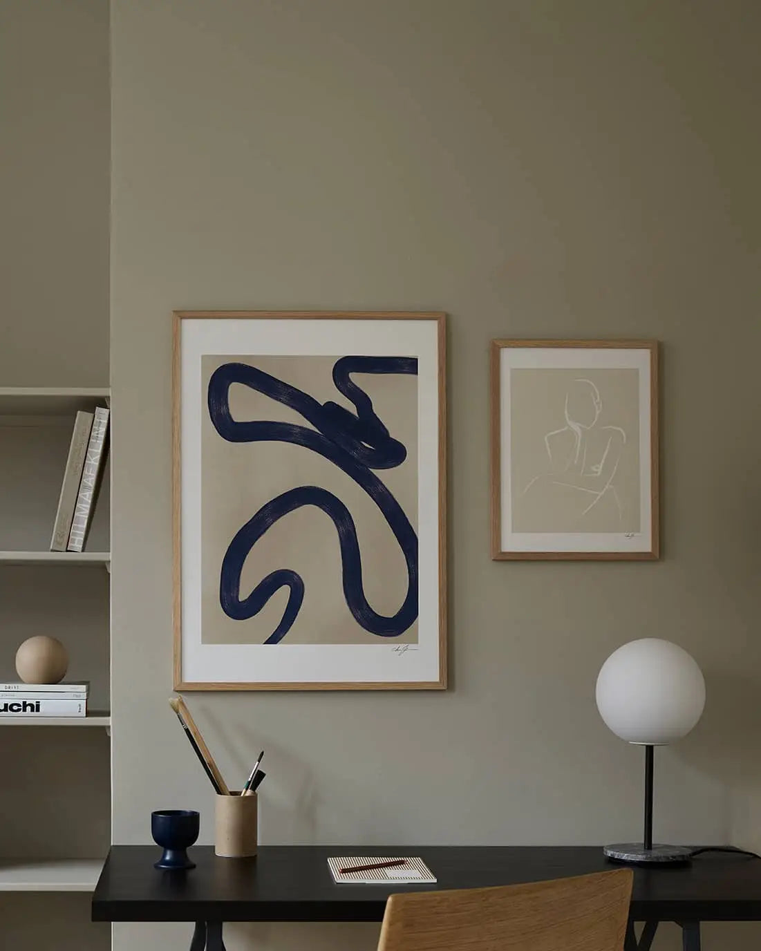 Minimalist workspace with a black desk, wooden chair, round white lamp, stationery, and abstract wall art in wooden frames; shelves with books and décor to the left feature The Poster Club Ana Frois - Orange Pendant (50x70) fine art print.