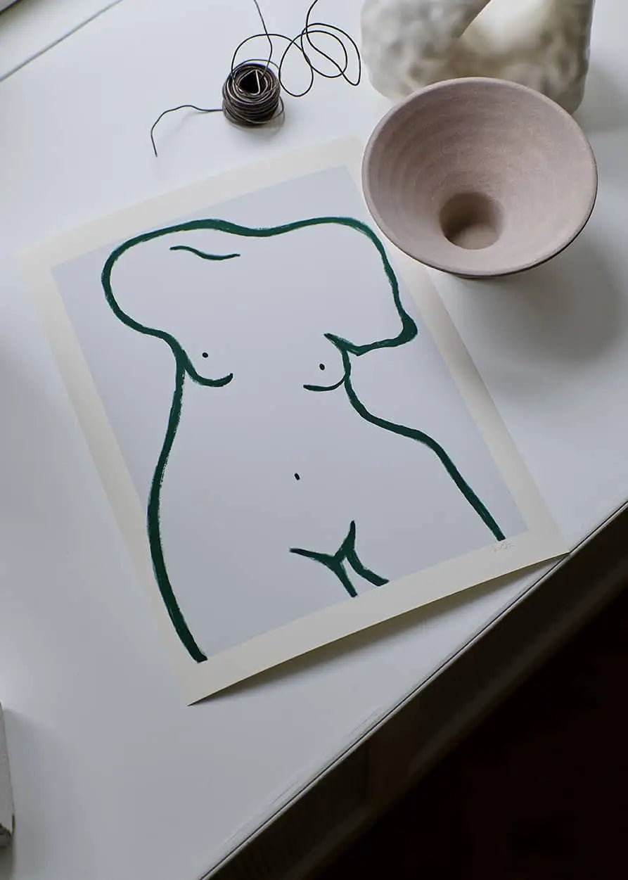 The Poster Club Anna Mörner - Statue (50x70) features a minimalist line drawing of a female torso on premium art paper, displayed on a white table with a pink ceramic bowl, wire coil, and blurred background object.