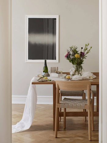 A wooden dining table with wine, glasses, flowers in a vase, food, and a white tablecloth is paired with two wooden chairs. The Poster Club’s Anne Nowak - Black Interstellar (30x40) art print completes the minimalist dining scene.