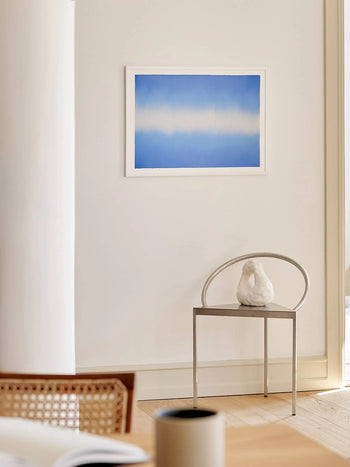 A minimalist room features a metal chair with a sculpted white object, set before a white wall displaying Anne Nowak - Blue Interstellar (30x40) from The Poster Club. Light wood flooring and part of a woven chair are also visible.