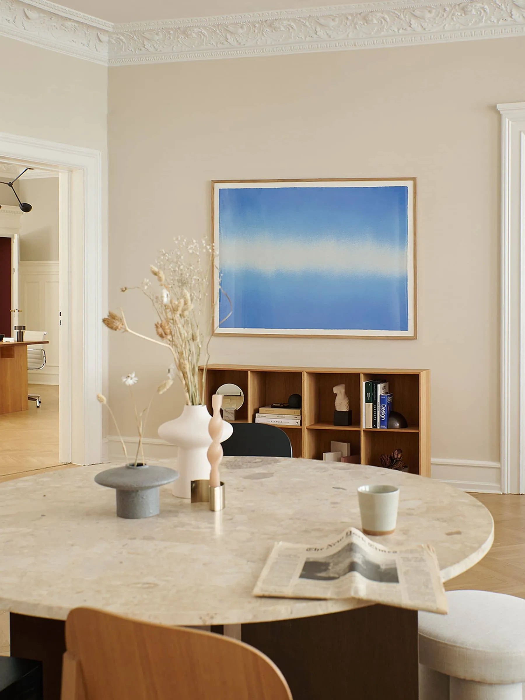 A round marble table with dried flowers, a cup, and a newspaper stands in a bright modern room. Anne Nowak’s Blue Interstellar (30x40) from The Poster Club hangs above a wooden shelf filled with books and decor.