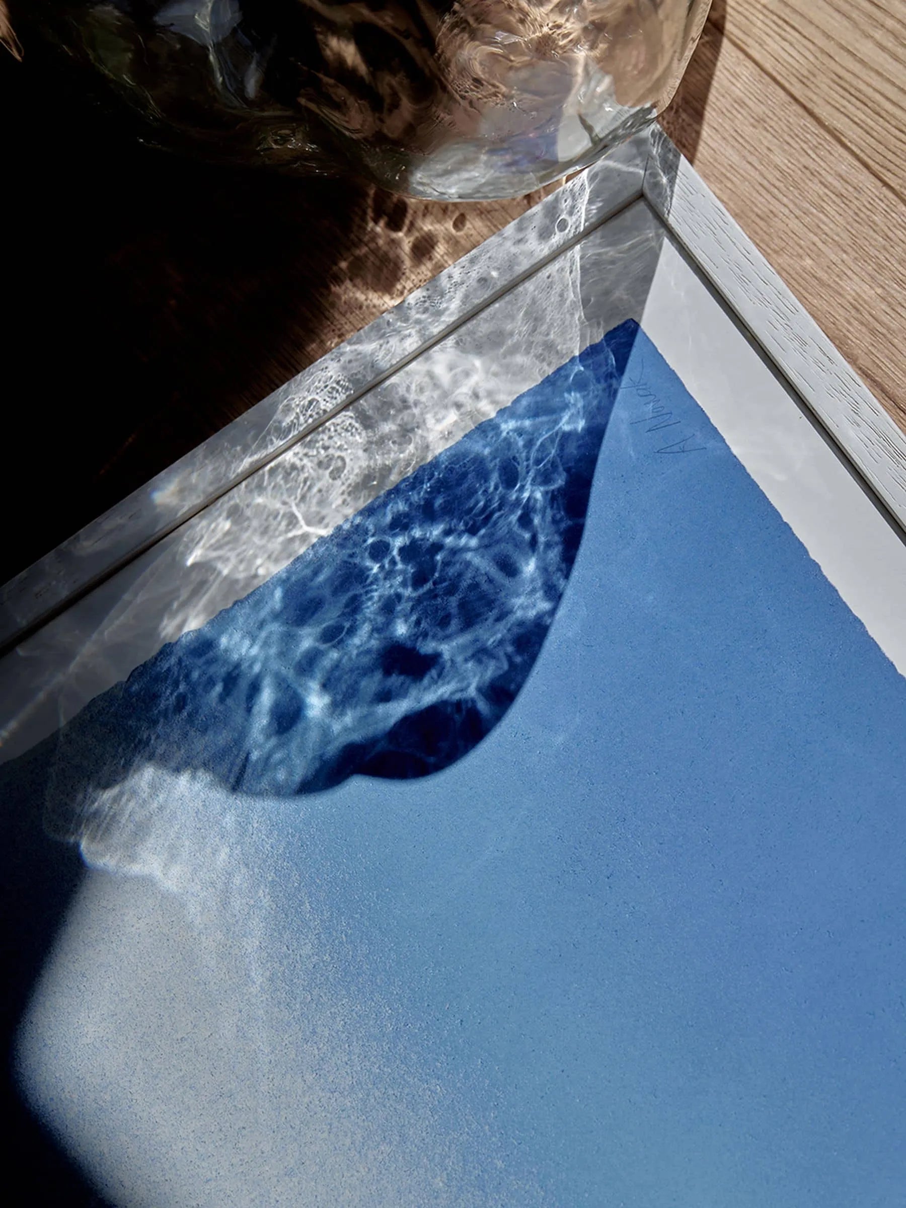 A framed Anne Nowak - Blue Interstellar (30x40) print from The Poster Club rests on a wooden surface, illuminated by sunlight. Its shimmering blue tones reflect Anne Nowak’s signature ethereal style.