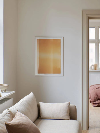A minimal living room with a light beige sofa and cushions, a white wall displaying Anne Nowak - Yellow Interstellar (30x40) from The Poster Club, and a view into a cozy bedroom with mauve bedding and sunlight from the window.